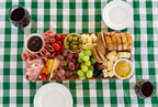 A charcuterie board arranged on a green and white checkered tablecloth, featuring sliced bread, cheeses, grapes, olives, crackers, salami, prosciutto, tomatoes, and small bowls of dips. Two glasses of red wine and two empty plates are also visible around the board.
